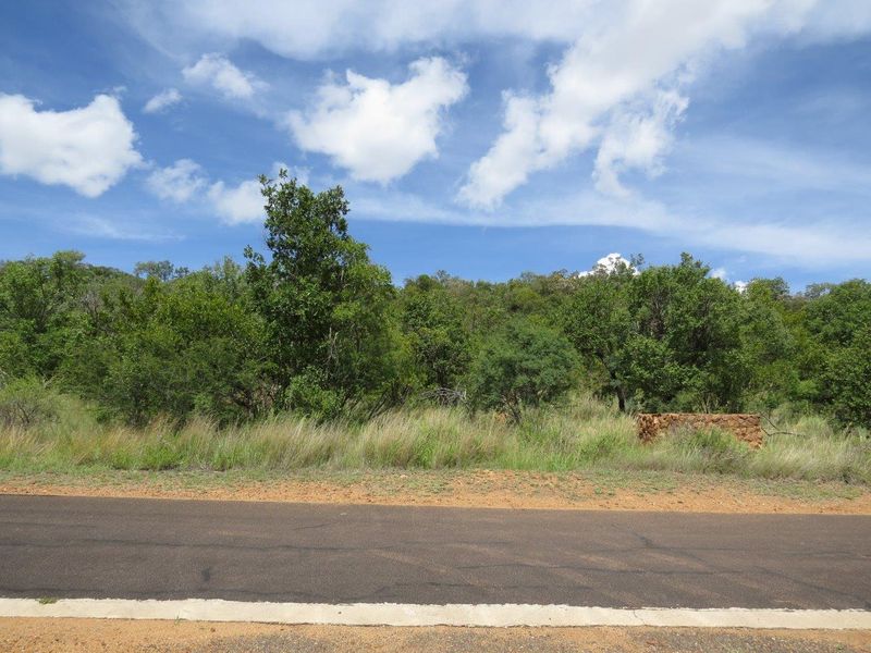 A view of the vacant residential land from the adjacent asphalt road, featuring natural vegetation and a stone boundary wall.