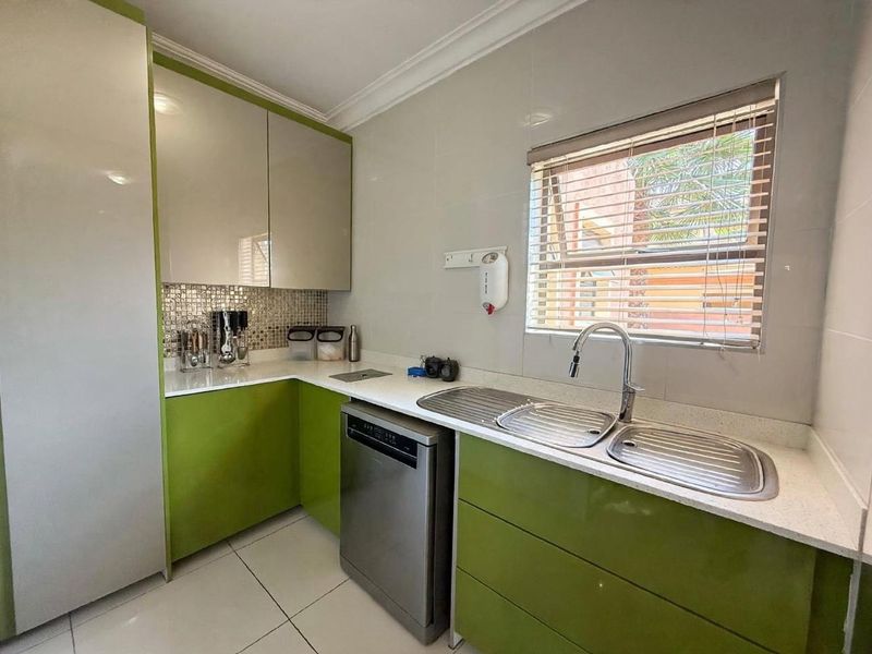 A modern scullery featuring vibrant green cabinetry, stone countertops, a double stainless steel sink, and tiled splashback with a window for natural light.