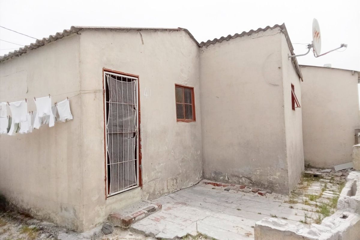 Rear exterior view of a modest dwelling featuring a security gate, satellite dish, and a paved area with visible signs of weathering and wear.
