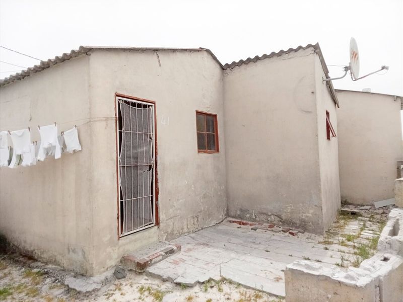 Rear exterior view of a modest dwelling featuring a security gate, satellite dish, and a paved area with visible signs of weathering and wear.