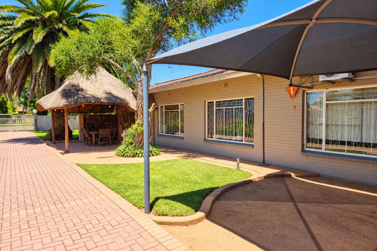 Rear exterior view featuring a paved driveway, a thatched lapa entertainment area, a shade sail, and a well-maintained garden space.