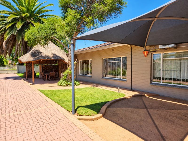 Rear exterior view featuring a paved driveway, a thatched lapa entertainment area, a shade sail, and a well-maintained garden space.