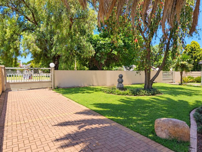 A well-maintained front garden featuring a paved driveway, established trees, and a secure boundary wall with a sliding gate.