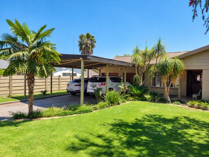 Front elevation of the property featuring a spacious carport, concrete driveway, and a well-maintained garden with established landscaping and boundary walling.