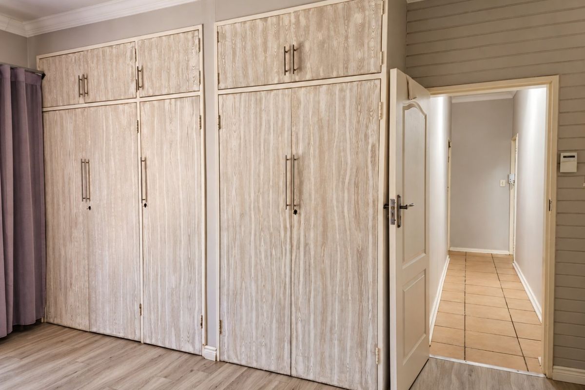 A clean bedroom featuring extensive built-in cupboards with wood-grain finish, laminate flooring, and neutral wall tones, leading into a tiled hallway.