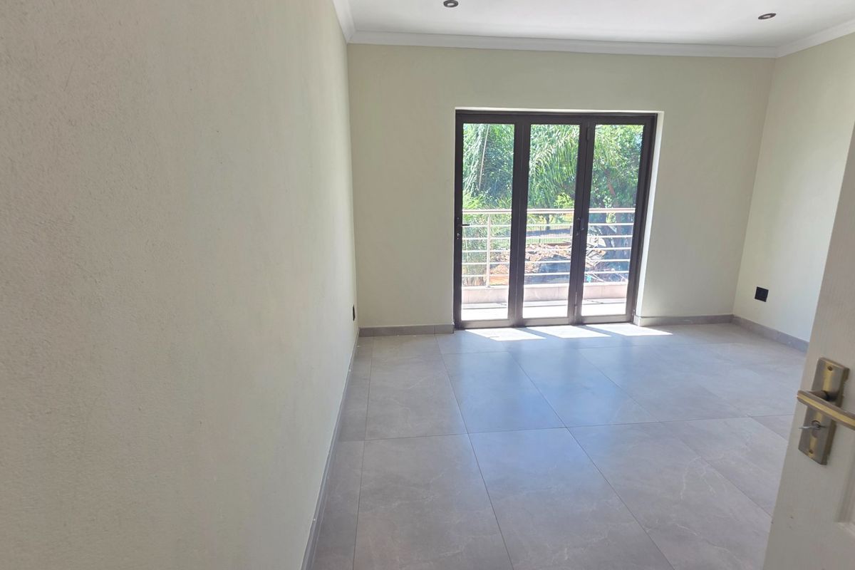 A bright, empty bedroom featuring modern large-format floor tiles, recessed lighting, and a sliding glass door providing access to an outdoor balcony.