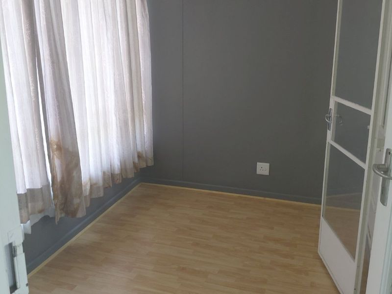 An empty bedroom featuring light-toned laminate flooring, grey painted walls, and a large window with curtains.