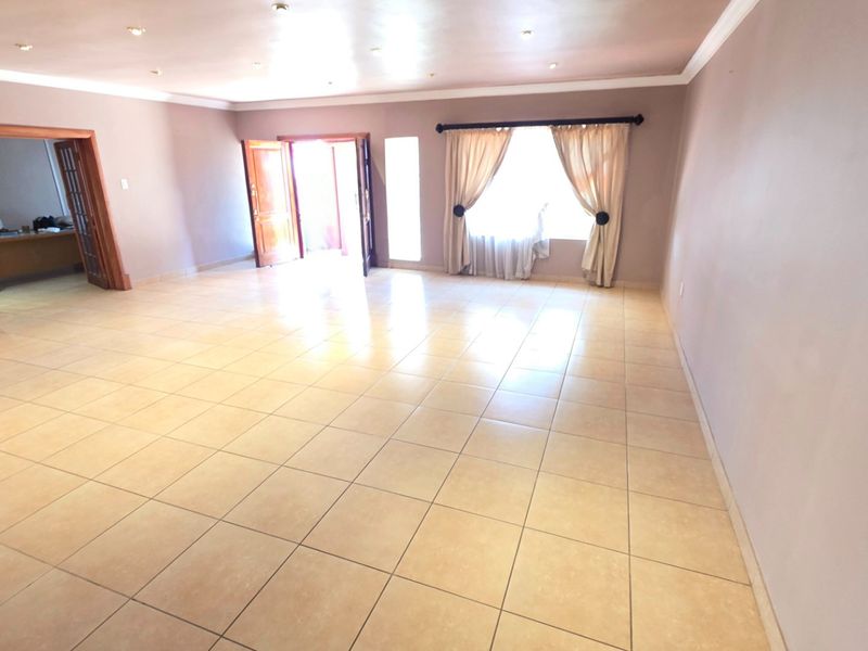 A spacious, empty living room featuring neutral tiled flooring, recessed downlighting, and natural light through a large window and double door entrance.