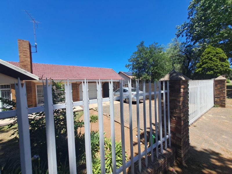 Front view of a single-story brick house featuring a tiled roof, chimney, paved driveway, and secure metal spike fencing with brick pillars.