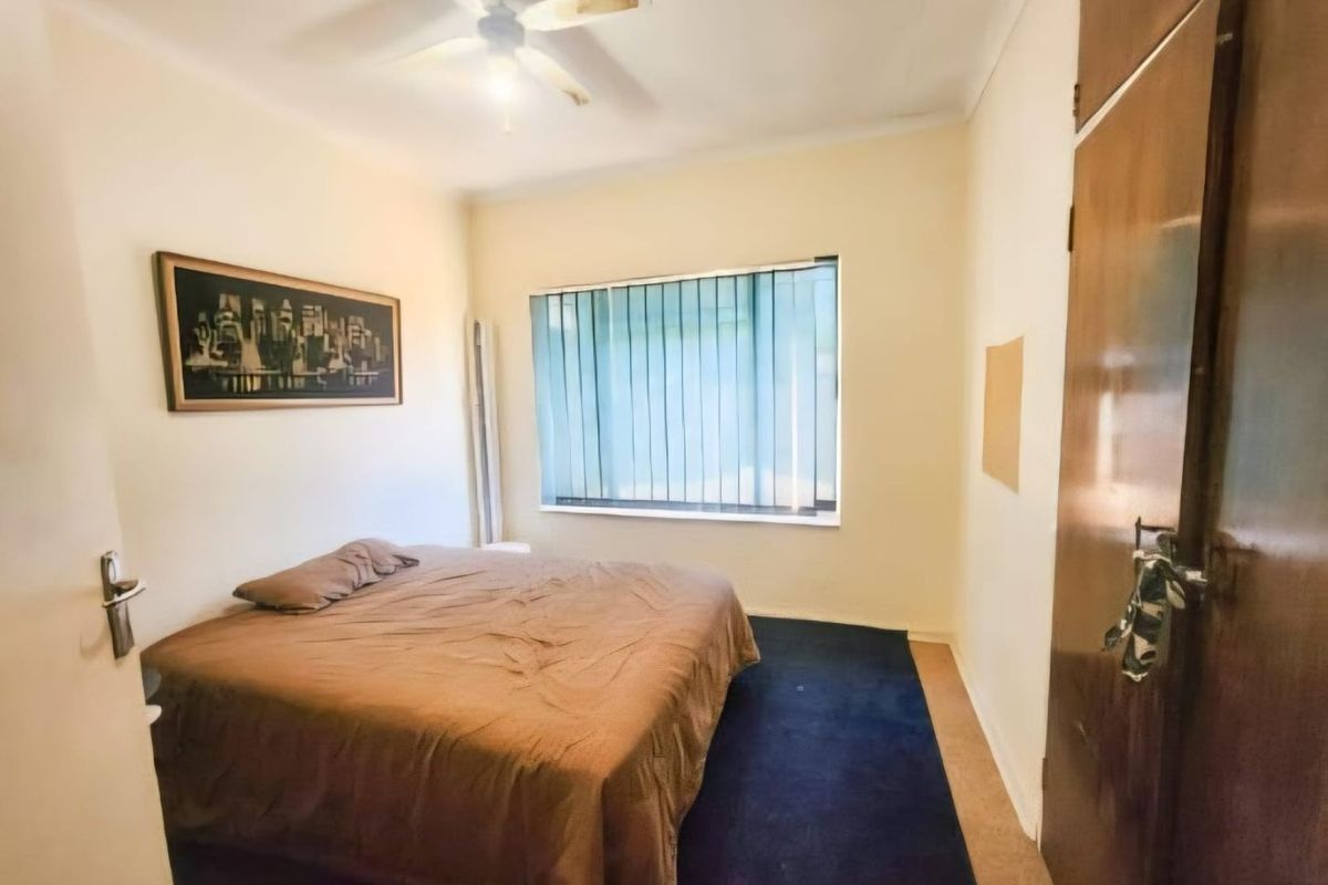 A bedroom featuring built-in wooden cupboards, a ceiling fan, and vertical blinds, with carpeted flooring.