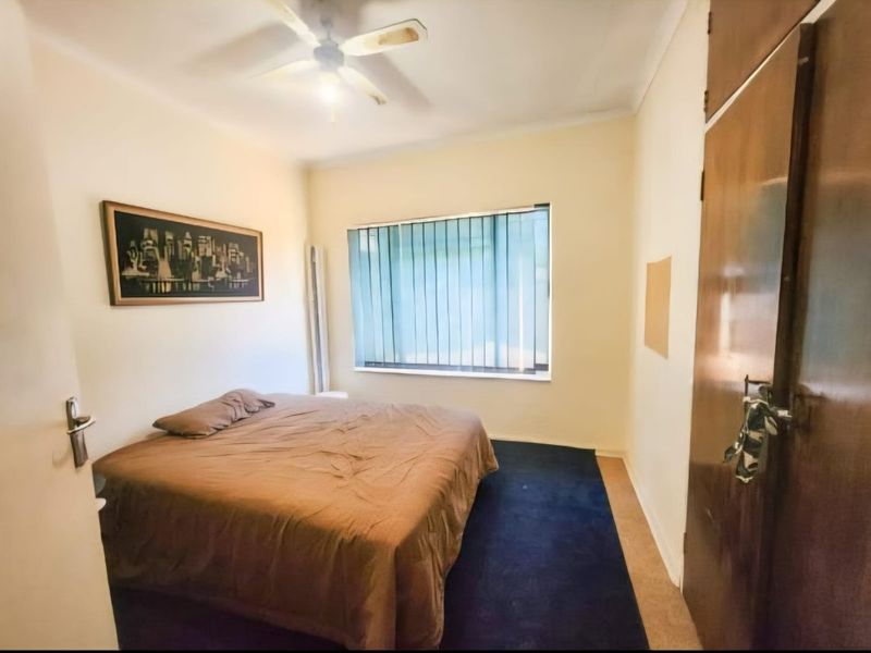 A bedroom featuring built-in wooden cupboards, a ceiling fan, and vertical blinds, with carpeted flooring.