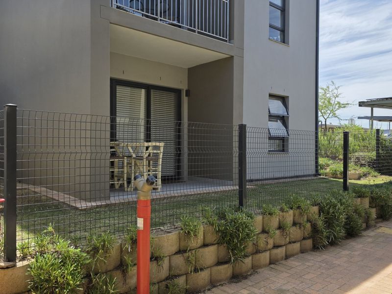 Exterior view of an apartment unit featuring a private patio, mesh fencing, and a landscaped retaining wall with a paved walkway.