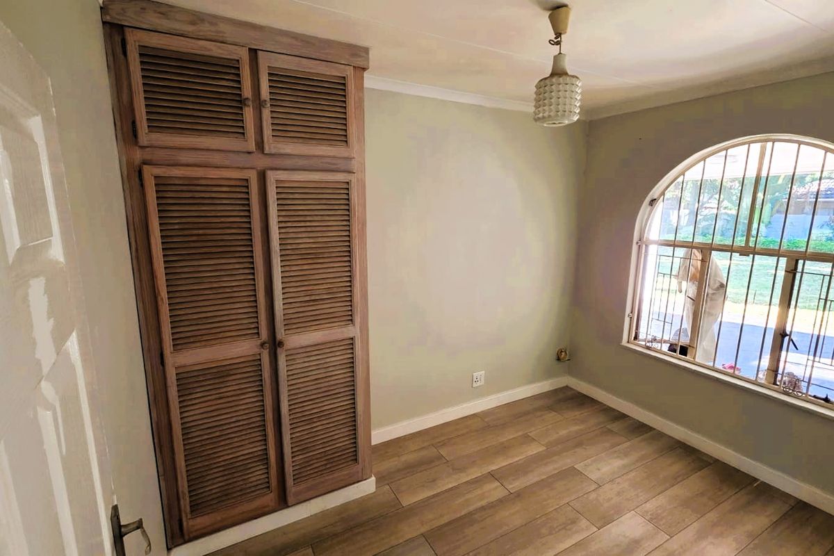A bright, empty bedroom featuring wood-look tile flooring, a built-in louvered wardrobe, and an arched window with security bars.