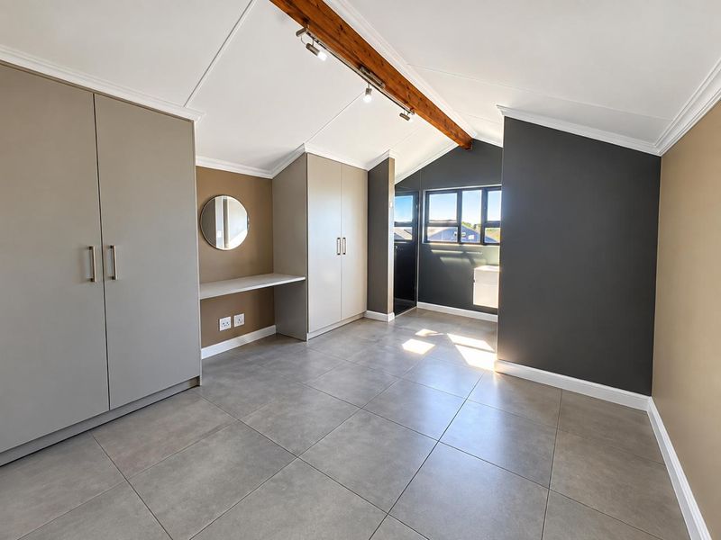 A modern, well-maintained bedroom featuring vaulted ceilings with exposed timber beams, built-in cupboards, a built-in desk, and large-format floor tiles.