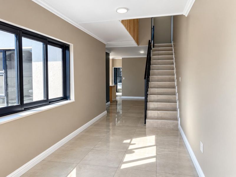 A bright, modern hallway featuring neutral walls, large format floor tiles, and a staircase with a sleek black metal balustrade.