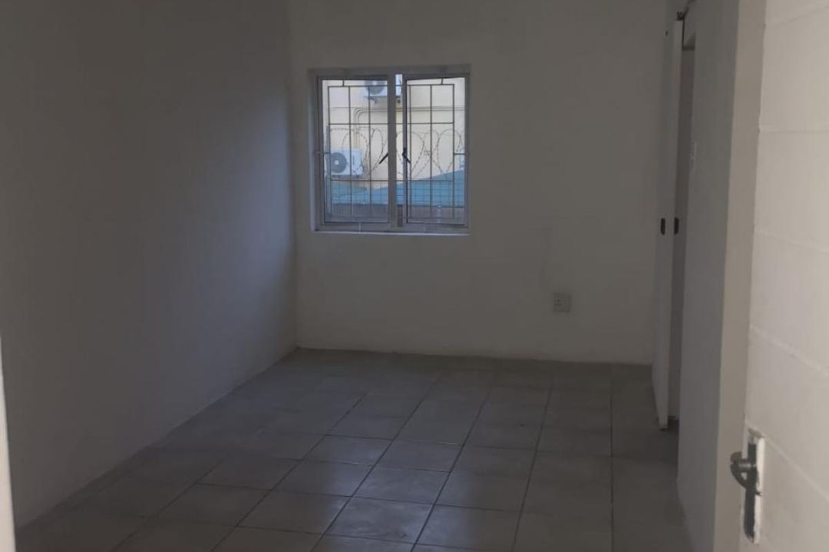 A vacant, neutral-toned office space featuring tiled flooring, a window with security bars, and basic ceiling fixtures, suitable for various commercial uses.