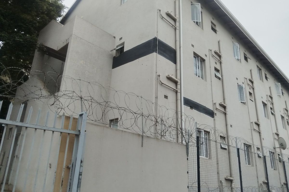 Exterior view of a multi-story commercial building featuring external plumbing, casement windows, and perimeter security fencing with razor wire.