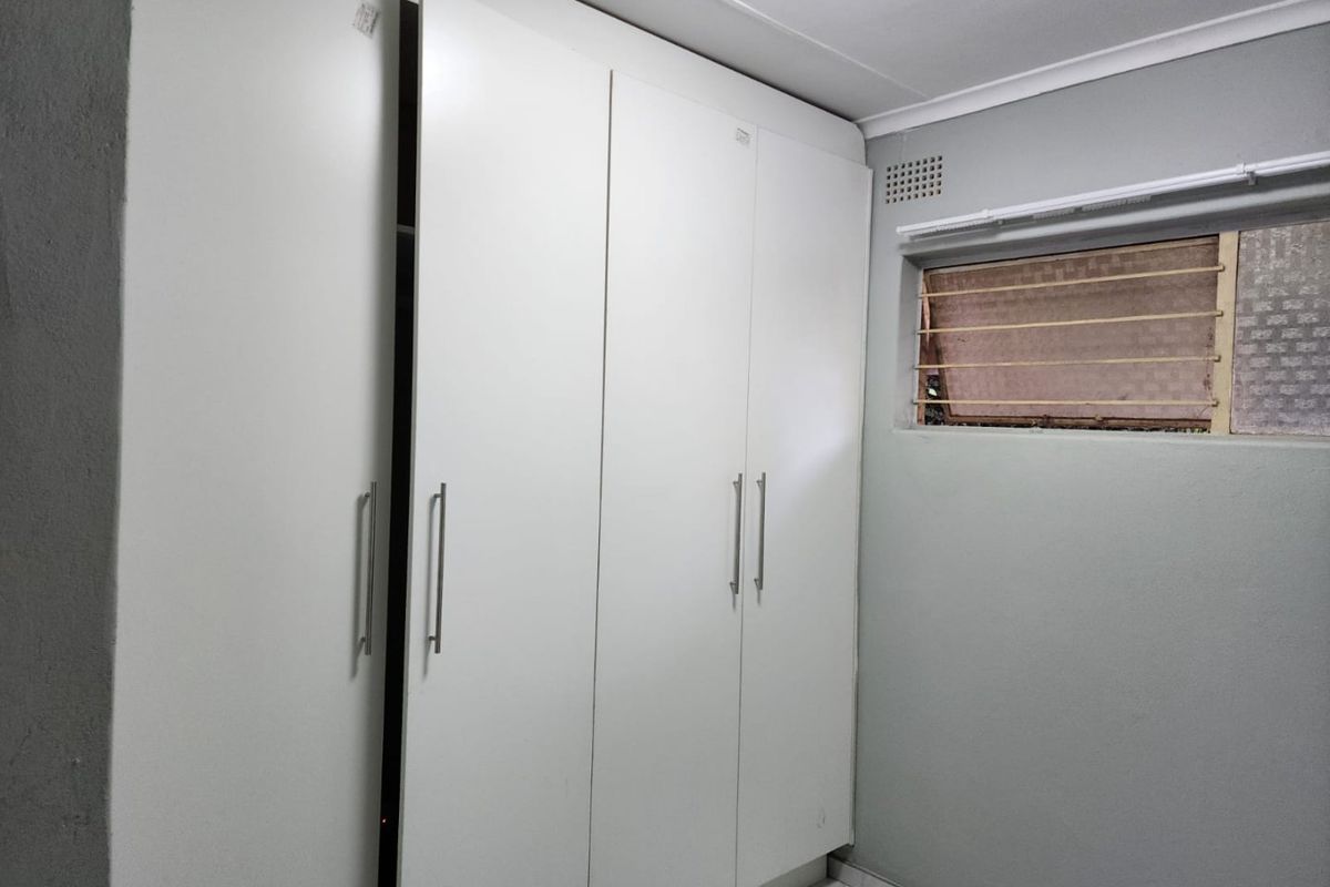 A bedroom featuring built-in cupboards, tiled flooring, and a top-hung window with security bars.