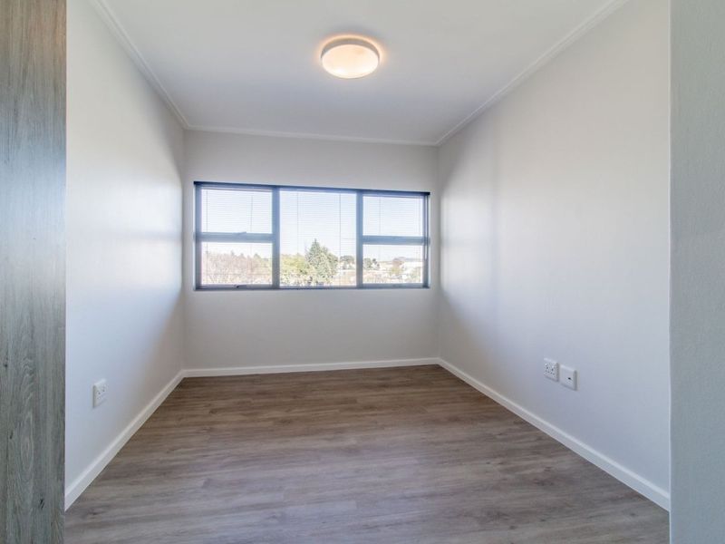 A bright, empty bedroom featuring modern laminate flooring, neutral walls, and large windows with fitted blinds, offering a clean and versatile living space.