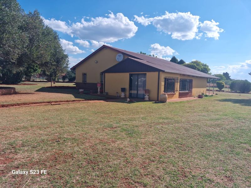Rear view of the farm house featuring a pitched roof, sliding glass doors, and security bars on the windows, set within a large garden.