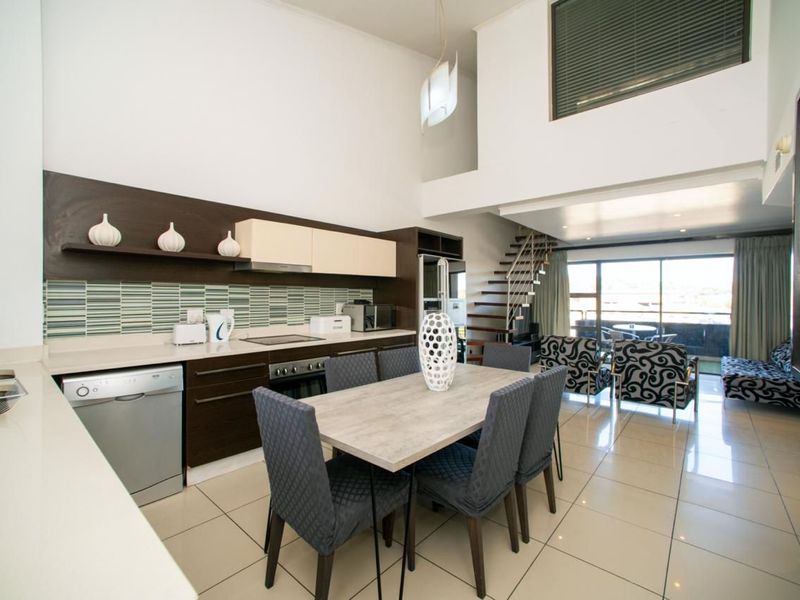 Spacious open-plan kitchen and dining area featuring modern cabinetry, stone countertops, tiled floors, and a staircase leading to a mezzanine level.