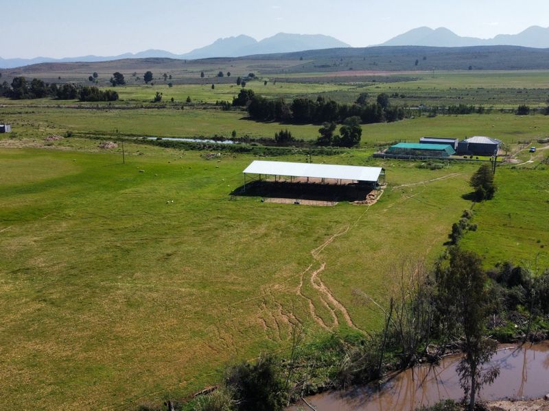 Aerial view of a farm property featuring open-sided agricultural structures, expansive grazing paddocks, and river frontage set against a mountain backdrop.