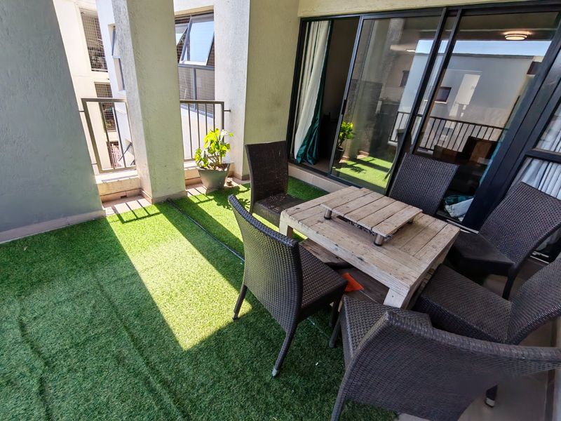 A covered balcony featuring artificial grass flooring, concrete structural pillars, and large sliding glass doors providing access to the interior living space.