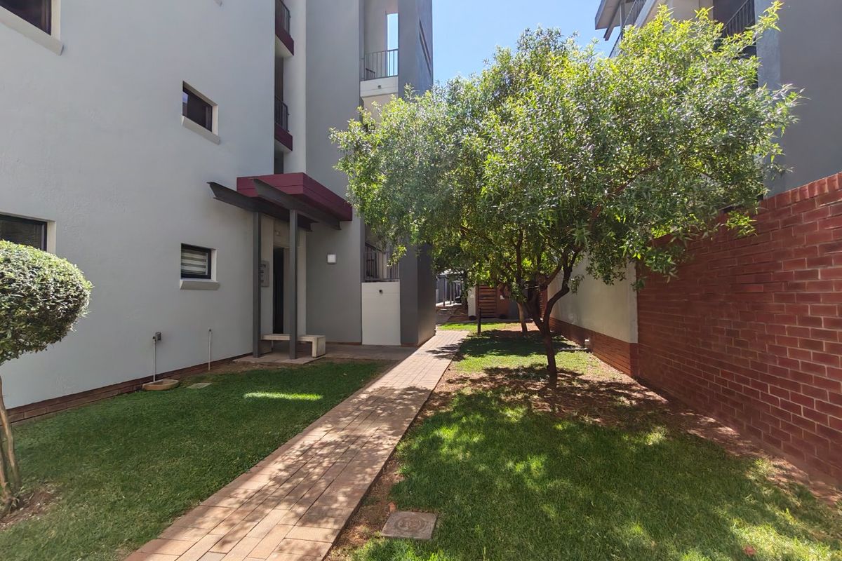 A well-maintained exterior rear garden area featuring a paved walkway, landscaped lawn, and a brick boundary wall alongside the apartment building facade.