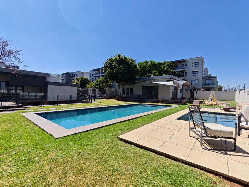 A well-maintained outdoor pool area featuring a rectangular swimming pool, paved decking, and landscaped lawn within a secure apartment complex setting.