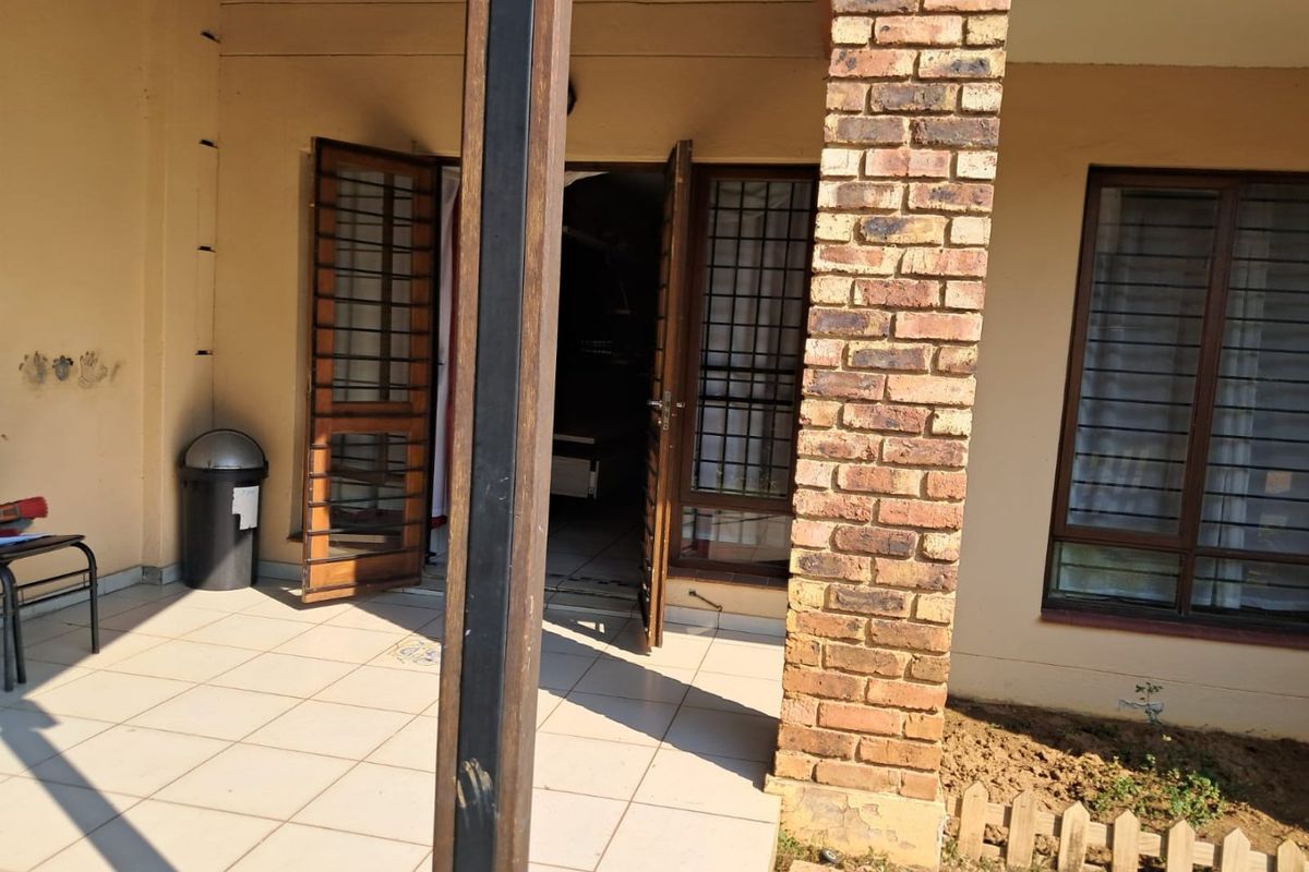 A covered patio area featuring tiled flooring, a brick support pillar, and secure timber-framed doors leading into the apartment.