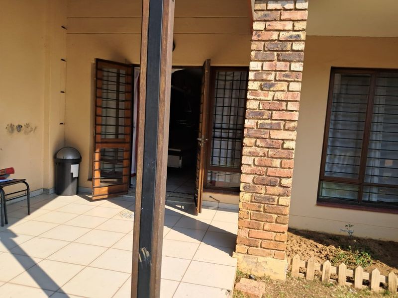 A covered patio area featuring tiled flooring, a brick support pillar, and secure timber-framed doors leading into the apartment.
