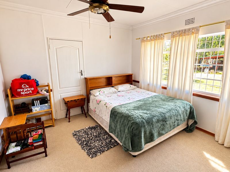 A well-lit bedroom featuring built-in cupboards, a ceiling fan, and large windows that provide natural light and views of the garden.