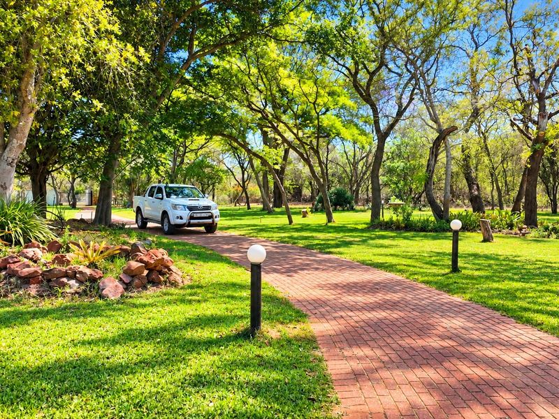 A well-maintained garden featuring a brick-paved pathway lined with bollard lights, surrounded by lush green lawns and mature trees on a farm property.