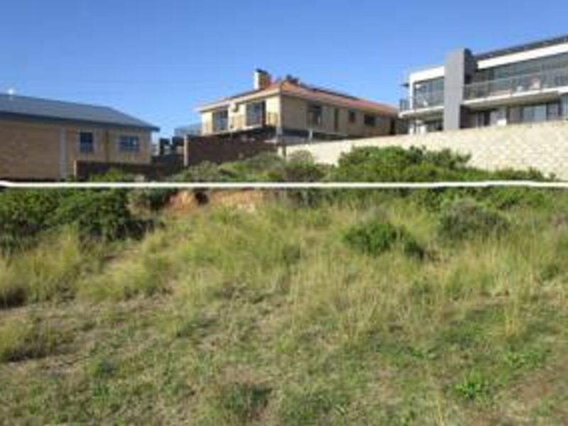 A vacant residential plot featuring natural vegetation, a retaining wall, and surrounding residential properties.