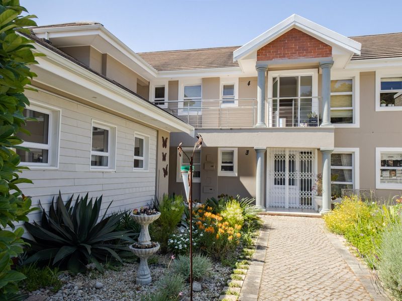 A well-maintained apartment complex entrance featuring a paved walkway, landscaped garden, and a balcony with stainless steel balustrades.
