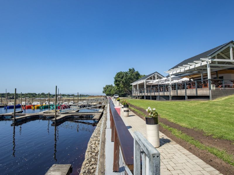 A scenic view of a waterfront property featuring a paved walkway, timber decking, and a pergola overlooking a private boat jetty and marina.