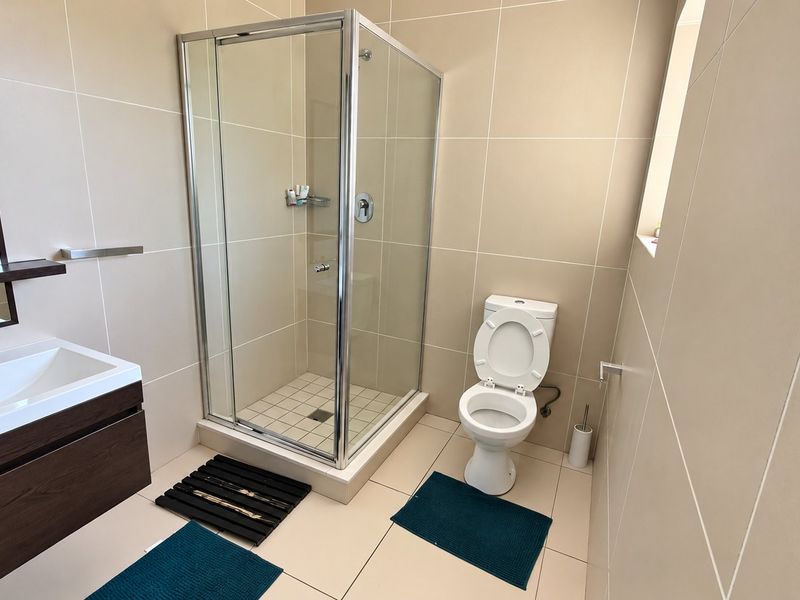 A modern, well-maintained bathroom featuring a glass shower enclosure, wall-mounted vanity with basin, and a standard toilet with neutral wall and floor tiling.