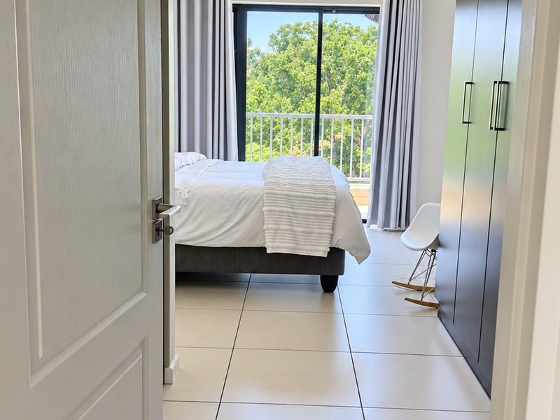A bright bedroom featuring large built-in cupboards, neutral tiled flooring, and a sliding glass door that opens onto a private balcony.