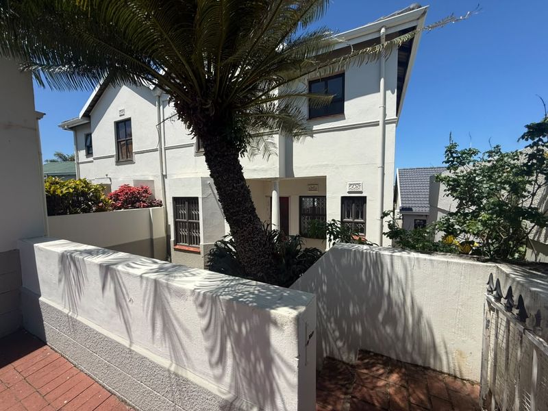 Front exterior view of a double-story townhouse featuring a paved entrance, boundary wall, and established garden elements.