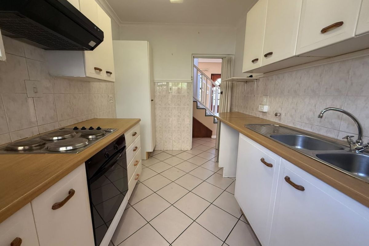 A functional galley-style kitchen featuring white cabinetry, laminate countertops, a built-in oven, electric hob, and a double stainless steel sink with tiled splashbacks.