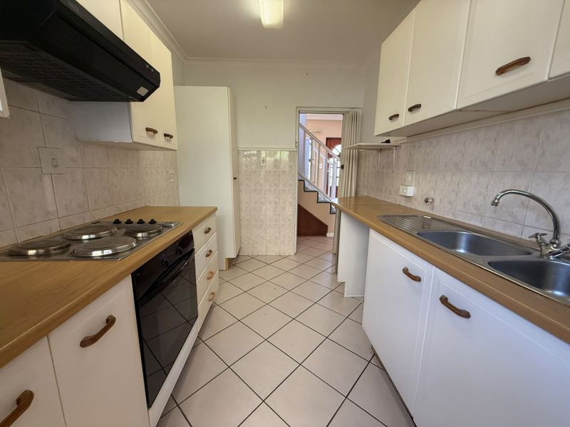 A functional galley-style kitchen featuring white cabinetry, laminate countertops, a built-in oven, electric hob, and a double stainless steel sink with tiled splashbacks.