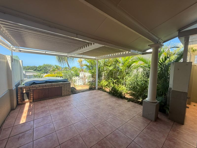 A covered outdoor patio featuring tiled flooring, a built-in brick braai, and a support column, overlooking a private garden area.