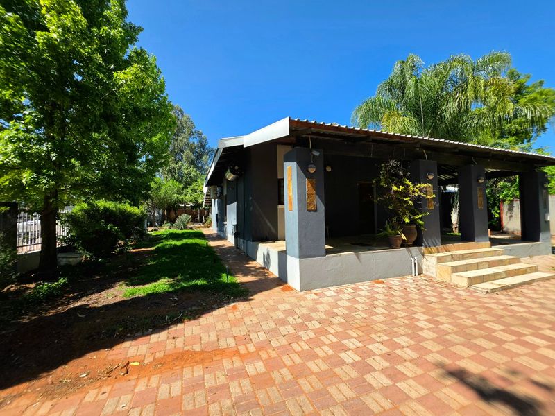 A rear exterior view featuring a covered patio with concrete pillars, tiled steps, and a brick-paved area set within a landscaped garden.