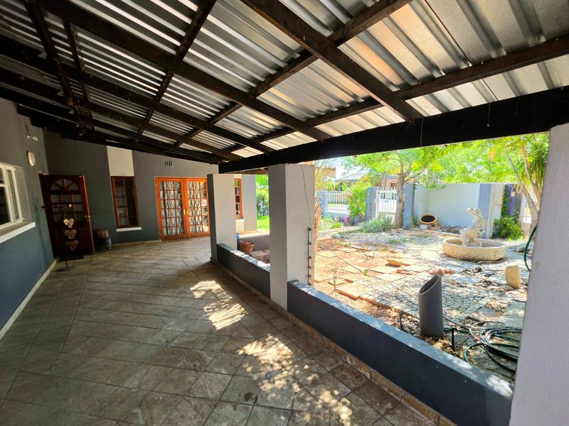 A covered outdoor patio area featuring tiled flooring, timber rafters, and a corrugated metal roof, overlooking a garden space with a stone fountain.
