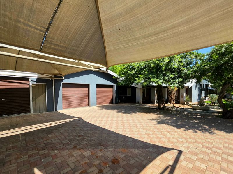 Front exterior view featuring a paved driveway, double garage with roller shutter doors, and a large overhead shade sail structure.
