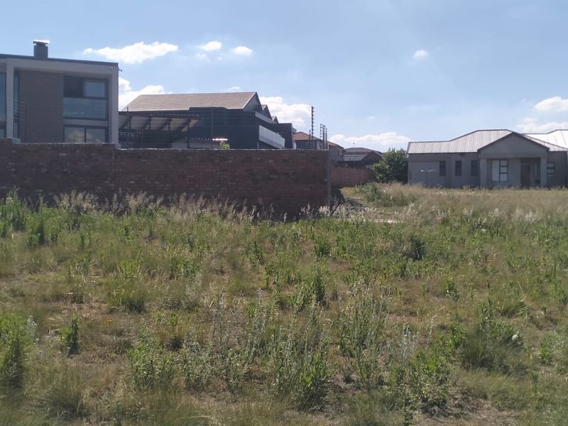 A vacant residential plot featuring natural grass coverage, enclosed by a brick boundary wall with integrated electric fencing in a developed neighborhood.