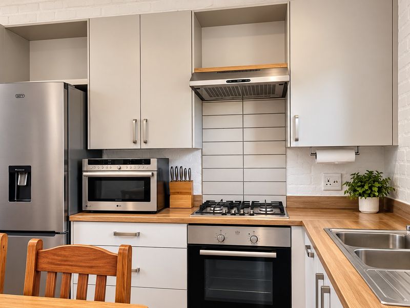 Modern kitchen featuring light grey cabinetry, wooden countertops, gas hob, electric oven, extractor fan, and a stainless steel sink against an exposed brick wall.