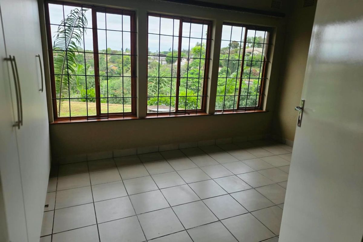 A bright, empty bedroom featuring tiled flooring, built-in cupboards, and large windows with security bars offering a pleasant garden view.