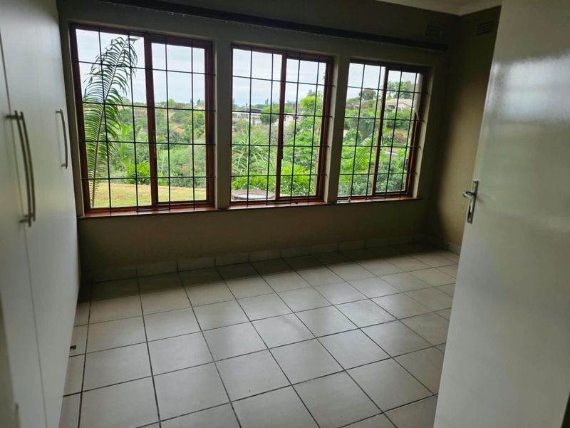 A bright, empty bedroom featuring tiled flooring, built-in cupboards, and large windows with security bars offering a pleasant garden view.