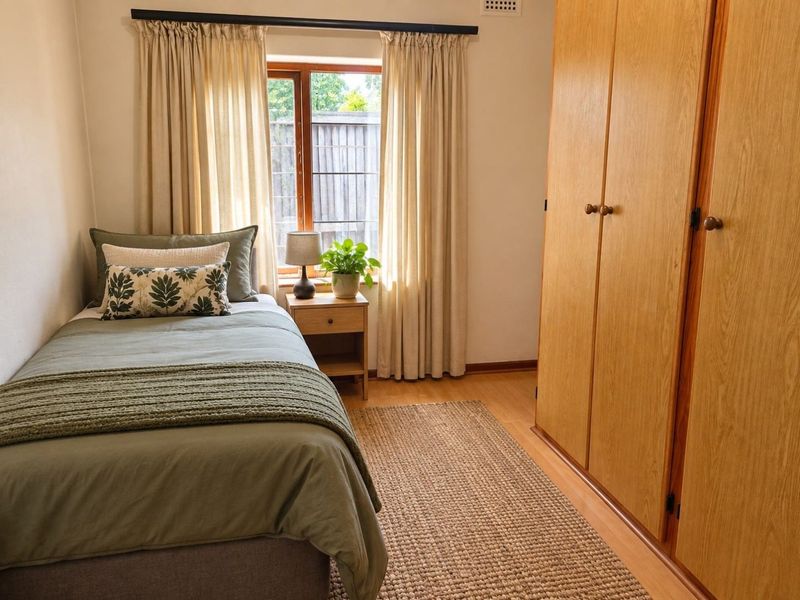 A cozy bedroom featuring built-in wooden wardrobes, laminate flooring, and natural light through a timber-framed window.